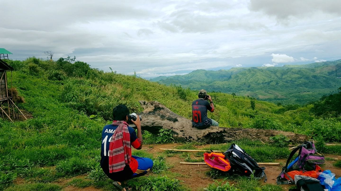 Two people crouched with cameras photographing a wide green valley from a hilltop, packs dropped beside them