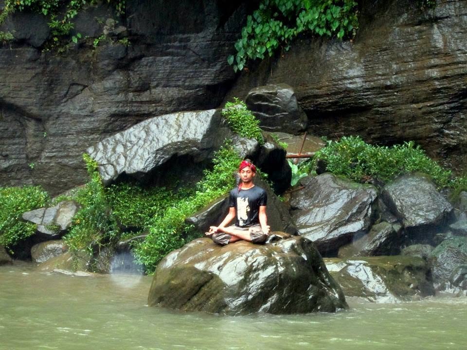 Sitting cross-legged on a boulder mid-river, cliff face and greenery rising behind