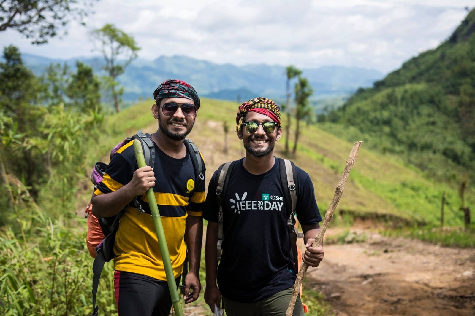 Two hikers with walking sticks and packs on a grassy hill trail, mountains in the distance