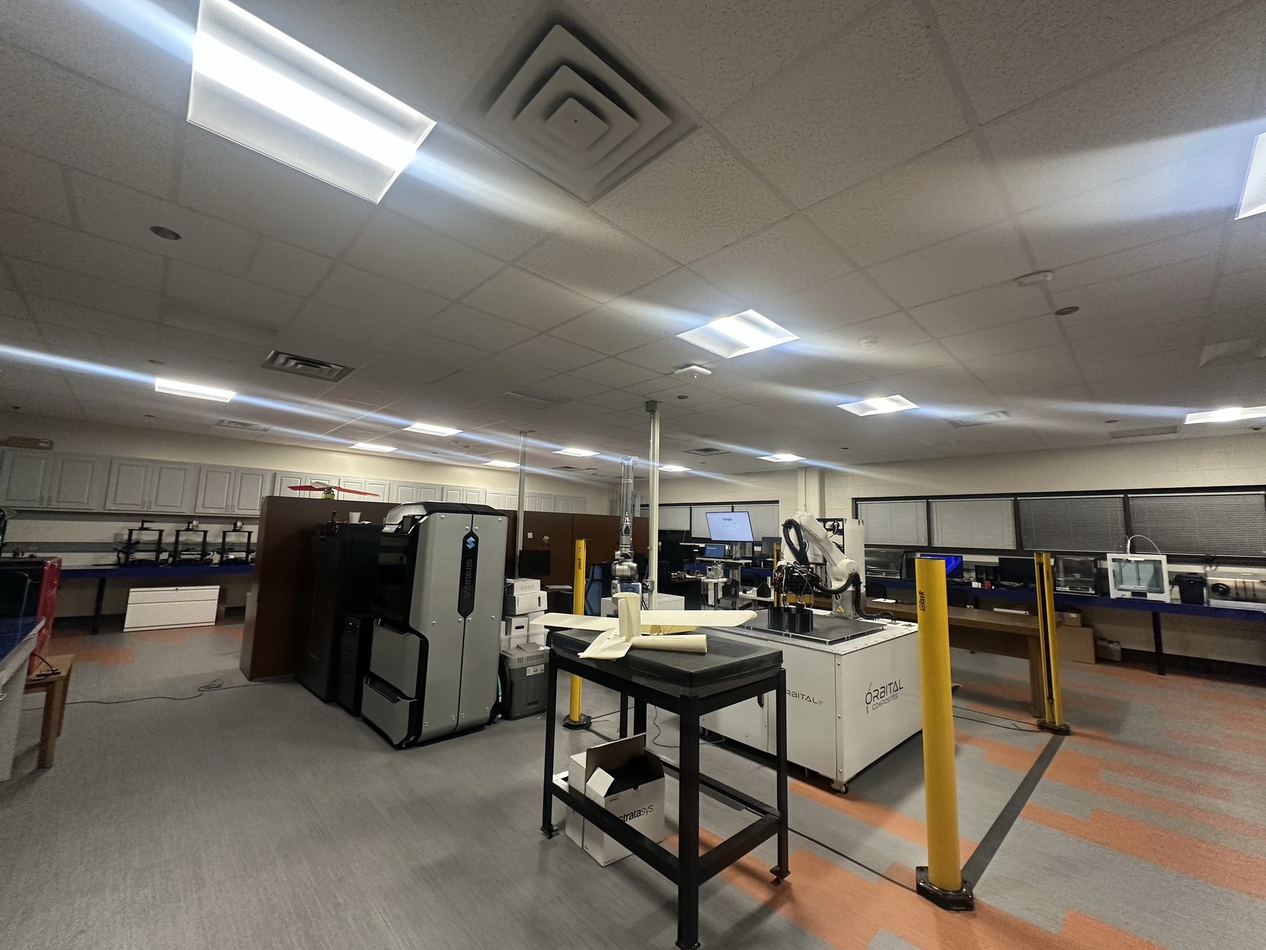 Additive manufacturing research laboratory at UT Arlington, with industrial and desktop FFF printers, a robotic composite deposition cell, and a printed UAV wing section